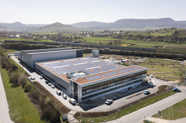 Luftbild einer großen Halle mit Solarmodulen auf dem Dach, umgeben von Fahrzeugen, Bäumen, Gras und Masten, mit Hügeln und einem klaren blauen Himmel im Hintergrund; die Halle ist Deutschlands erste solarbetriebene Fabrik.