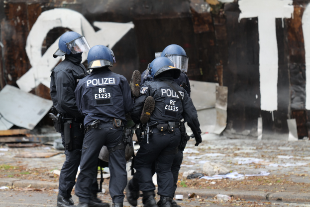 Eine Gruppe von Polizisten in schwarzen Uniformen und blauen Helmen steht vor einem Gebäude mit Graffiti, umgeben von verstreuten Papieren und Gegenständen auf dem Boden.