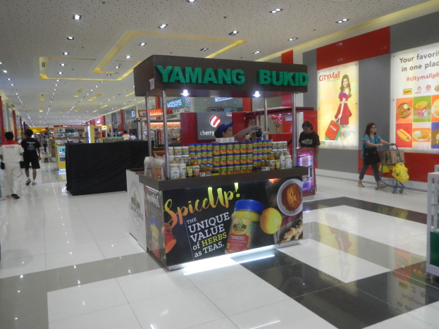 Ein belebtes Einkaufszentrum mit Menschen, die einkaufen, Taschen tragen und einen Stand mit Flaschen und anderen Artikeln, sowie Schilder mit Text und Bildern und einer Decke mit Lichtern.