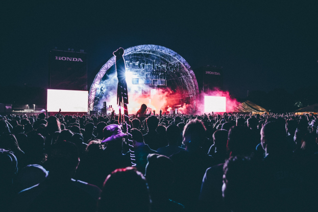 Eine große Menschenmenge auf einem Musikfestival mit einem Künstler auf einer beleuchteten Bühne umgeben von Musikern, Bildschirmen und Bannern in einer dunklen Umgebung.