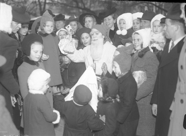Schwarzes und weißes Bild von Santa Claus, der zwischen Kindern mit Mützen steht, mit Bäumen im Hintergrund und Text unten.