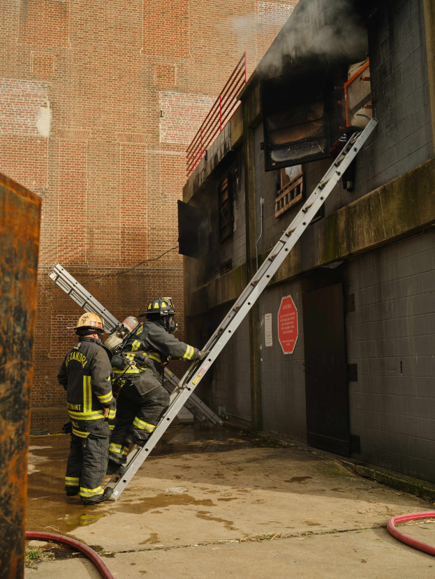 Feuerwehrleute mit Helmen klettern an einer Leiter zu einem raucherfüllten Gebäude hoch, während ein metallisches Objekt und Bodenschläuche sichtbar sind.