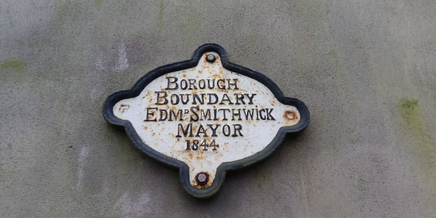 Metallplatte an einer Gebäudeseite mit schwarzem Hintergrund und weißer fetter Schrift, die "Borough Boundary Edm Smithwick Mayor" liest.