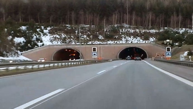 Autobahn mit Autos, die durch einen von Geländern und Texttafeln gesäumten Tunnel fahren, Schnee auf dem Boden, Bäume und Masten im Hintergrund und sichtbarer Himmel darüber.