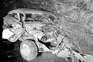 Ein Schwarz-Weiß-Foto von einem schwerbeschädigten Auto am Straßenrand mit einer zertrümmerten Front und zersplitterten Fenstern, einem Baum im Hintergrund und verstreuten Gegenständen.
