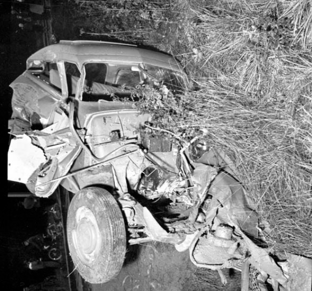Ein Schwarz-Weiß-Foto von einem schwerbeschädigten Auto am Straßenrand mit einer zertrümmerten Front und zersplitterten Fenstern, einem Baum im Hintergrund und verstreuten Gegenständen.