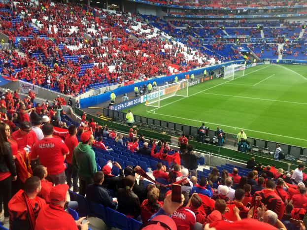 Ein volles Stadion mit sitzenden und stehenden Zuschauern, die ein Fußballspiel beobachten, mit sichtbaren Torpfosten, Bannern und Deckenleuchten unter einer Dachkonstruktion.
