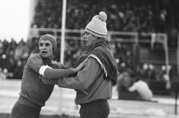 Zwei Männer in Jacken und Mützen auf einem Feld im Kampf mit unscharfen Zuschauern, Pfählen und einem Zaun im Hintergrund, in Schwarz-Weiß.