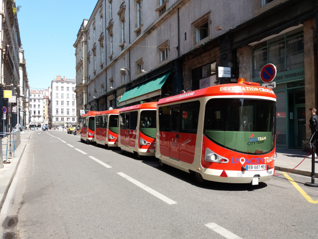 Eine Reihe roter und weißer Busse, die entlang einer Straße in Lyon, Frankreich, geparkt sind, mit Passanten auf dem benachbarten Gehweg und Gebäuden im Hintergrund.