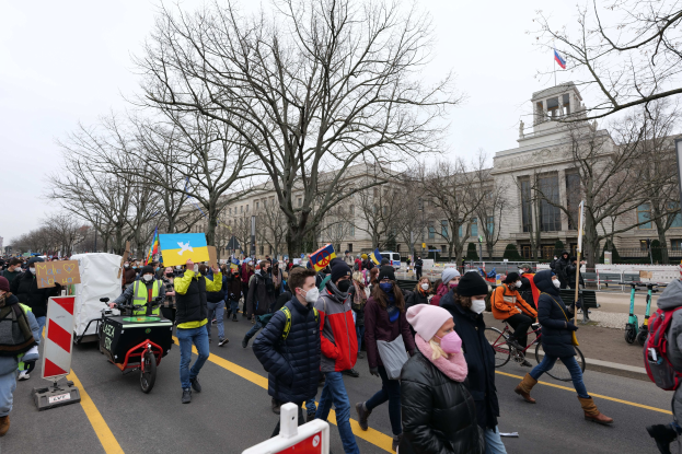 Eine große Protestmarsch mit Menschen, die Schilder und Plakate halten und Fahrräder fahren, geht an einem Gebäude in Washington, D.C. am 21. Januar 2020 vorbei, bei klarem blauem Himmel mit Bäumen und Schildern im Hintergrund.