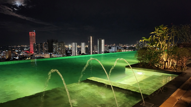 Ein Schwimmbad mit einem zentralen Brunnen, umgeben von Pflanzen auf der rechten Seite und Gebäuden im Hintergrund, beleuchtet von Stadtlichtern bei Nacht.