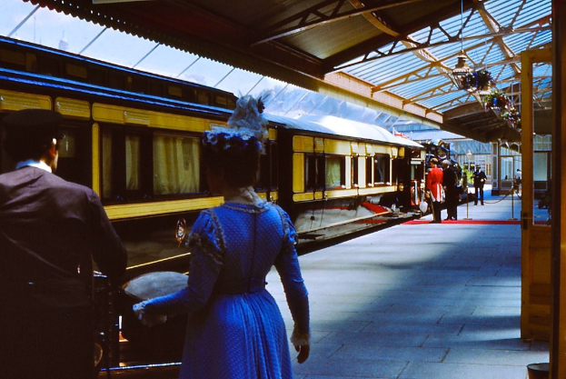 Frau in blauem Kleid neben einem Zug an einem Bahnhof mit Menschen auf dem Bahnsteig und dem Zug links.