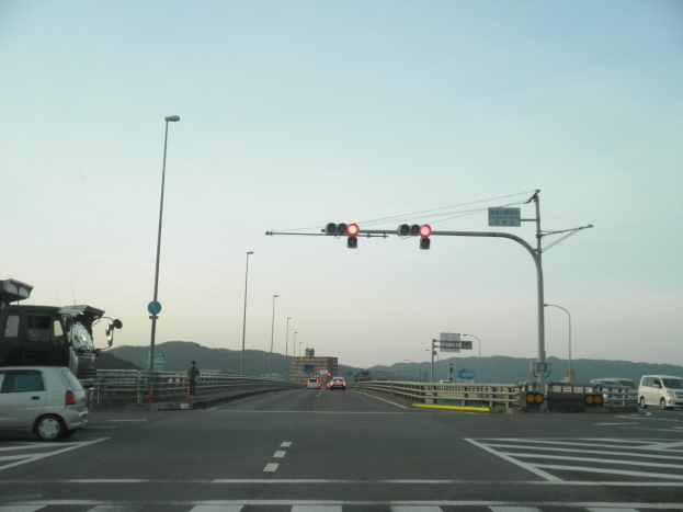 Ein Auto fährt auf einer Autobahn an einer Kreuzung mit Ampeln, umgeben von Fahrzeugen, einem Fußgänger auf dem Gehweg, Geländern, Laternen, Verkehrszeichen, Schildern, Gebäuden, Hügeln und einem klaren blauen Himmel.