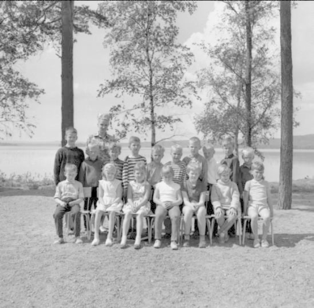 Schwarzes und weißes Foto von Kindern, die vor einem See posieren, mit Bäumen, Pflanzen, Hügeln, Wolken im Hintergrund und Text unten, der "1940er Kinder am See" lautet.