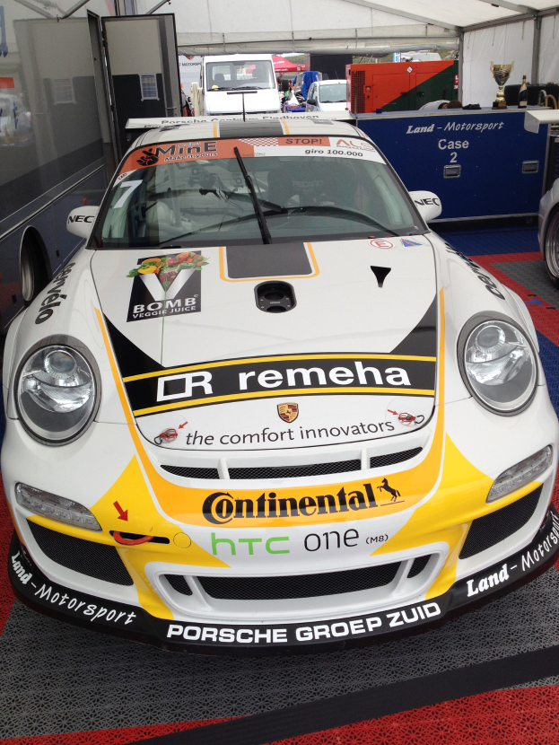 Weißer und gelber Porsche 911 GT3 RSR Rennwagen in einer Garage mit anderen Fahrzeugen, Menschen und Gegenständen geparkt.