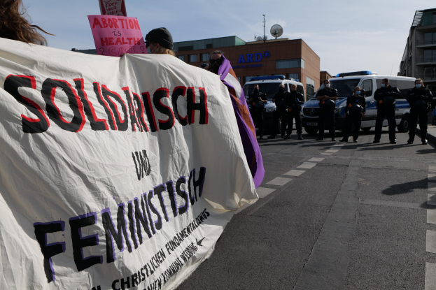 Gruppe von Menschen marschiert auf einer Straße und hält ein Banner mit der Aufschrift 'Solidarität und Feminismus', mit geparkten Fahrzeugen, Gebäuden, einer Schüsselantenne und einem klaren blauen Himmel im Hintergrund.