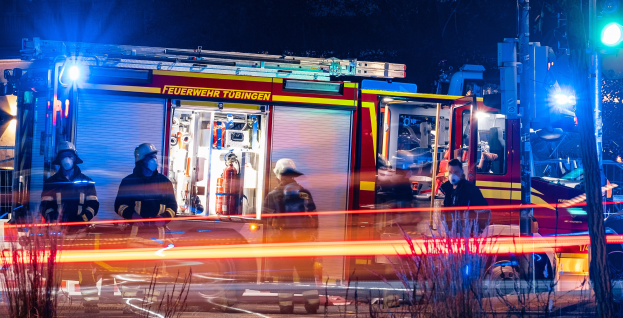 Feuerwehrleute in Schutzausrüstung vor einem beleuchteten Feuerwehrauto bei Nacht, mit Pflanzen im Vordergrund, einem Verkehrszeichen auf der rechten Seite und Bäumen im Hintergrund.
