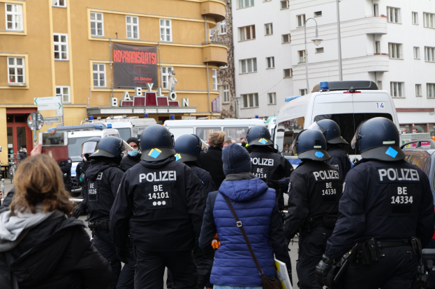 Polizeibeamte in Uniform vor einer Menge mit Helmen und Jacken während einer Demonstration in Berlin, Deutschland, mit Fahrzeugen, Gebäuden, Laternenmasten und einem Banner im Hintergrund.