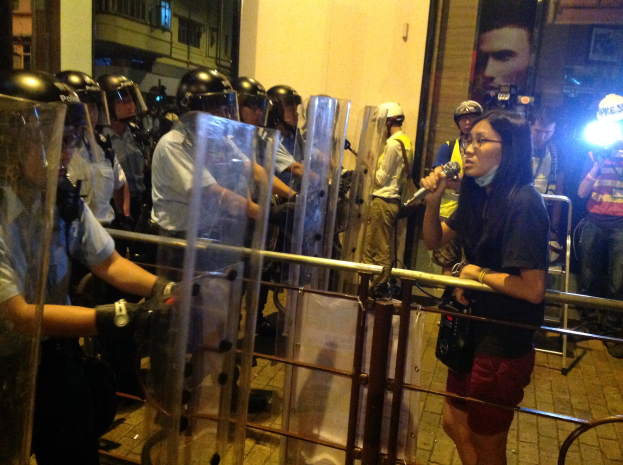 Eine Frau in einem schwarzen T-Shirt und roten Shorts steht vor der Bereitschaftspolizei, ein Mikrofon haltend, mit Menschen in Helmen und einer Metallabsperrung dahinter.