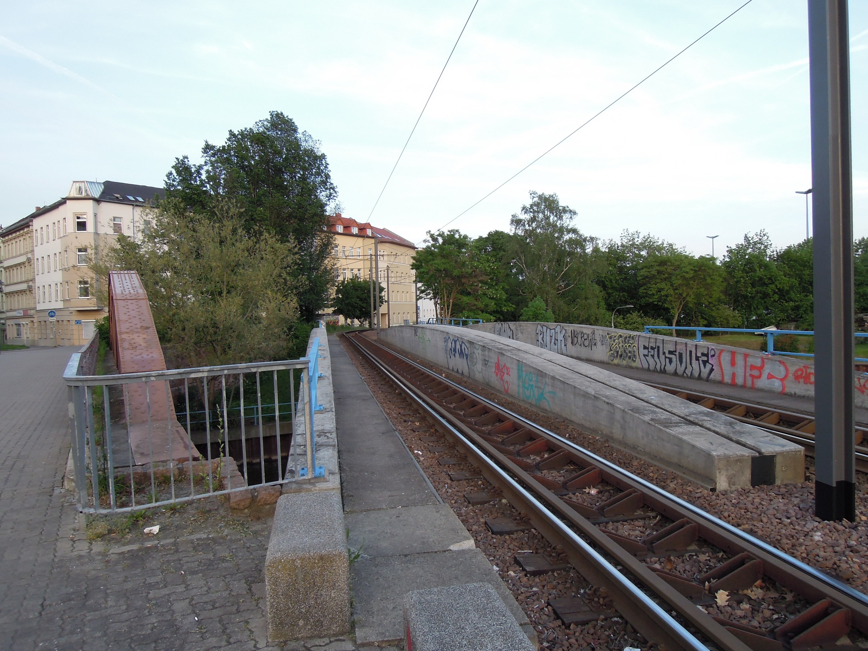 Bahnschiene mit Graffiti, umgeben von Steinen und einem Metallzaun, in der Nähe von Bäumen, Gebäuden und einem bewölkten Himmel.