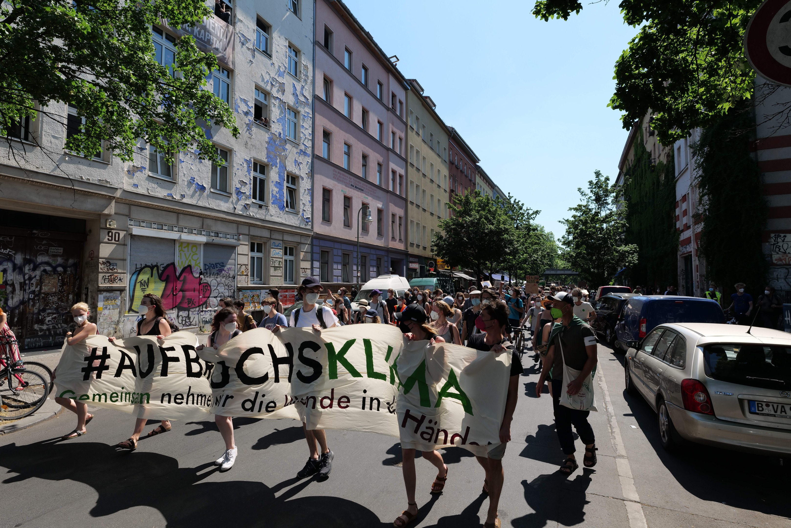 Eine Gruppe von Menschen, die eine Straße entlanggehen und ein Transparent halten, mit parkenden Autos auf der rechten Seite und einem Fahrradfahrer auf der linken Seite, vor Gebäuden und Bäumen während einer Demonstration in Berlin.