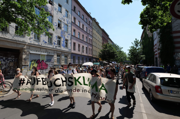 Eine Gruppe von Menschen, die eine Straße entlanggehen und ein Transparent halten, mit parkenden Autos auf der rechten Seite und einem Fahrradfahrer auf der linken Seite, vor Gebäuden und Bäumen während einer Demonstration in Berlin.