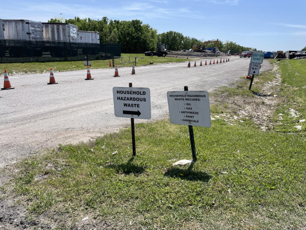 Straßensammelstelle für "Haushaltsgefährliche Abfälle" markiert durch Verkehrskegel, mit Fahrzeugen, Gras, Bäumen und bewölktem Himmel im Hintergrund.