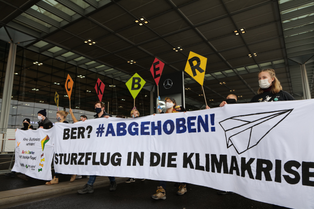 Eine Gruppe maskierter Individuals mit einem Banner 'Sturzflug in die Klimakrise' vor einem Glasgebäude mit Säulen, beleuchtet von Deckenlampen.