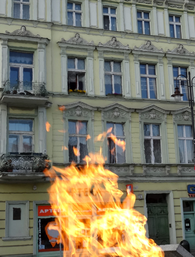 Ein großes Feuer brennt vor einem Gebäude mit Fenstern, Türen, einem Laternenmast und einem Schild.
