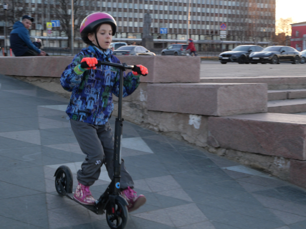 Ein Junge mit Helm und Handschuhen fährt auf einem Gehweg mit Treppen, Fahrzeugen, Menschen, Bäumen, Pfählen, Brettern, Gebäuden und einem klaren blauen Himmel im Hintergrund.