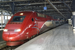 Ein roter Hochgeschwindigkeitszug steht auf einem Bahnhof mit einer Plattform auf der rechten Seite, einige Menschen stehen herum und im Hintergrund sind Säulen, Stangen und andere Gegenstände zu sehen.