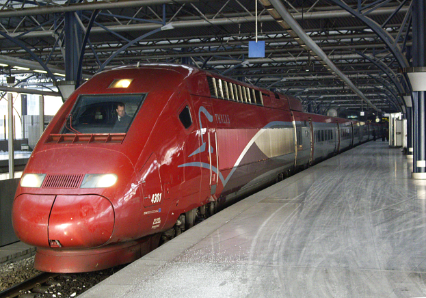 Ein roter Hochgeschwindigkeitszug steht auf einem Bahnhof mit einer Plattform auf der rechten Seite, einige Menschen stehen herum und im Hintergrund sind Säulen, Stangen und andere Gegenstände zu sehen.