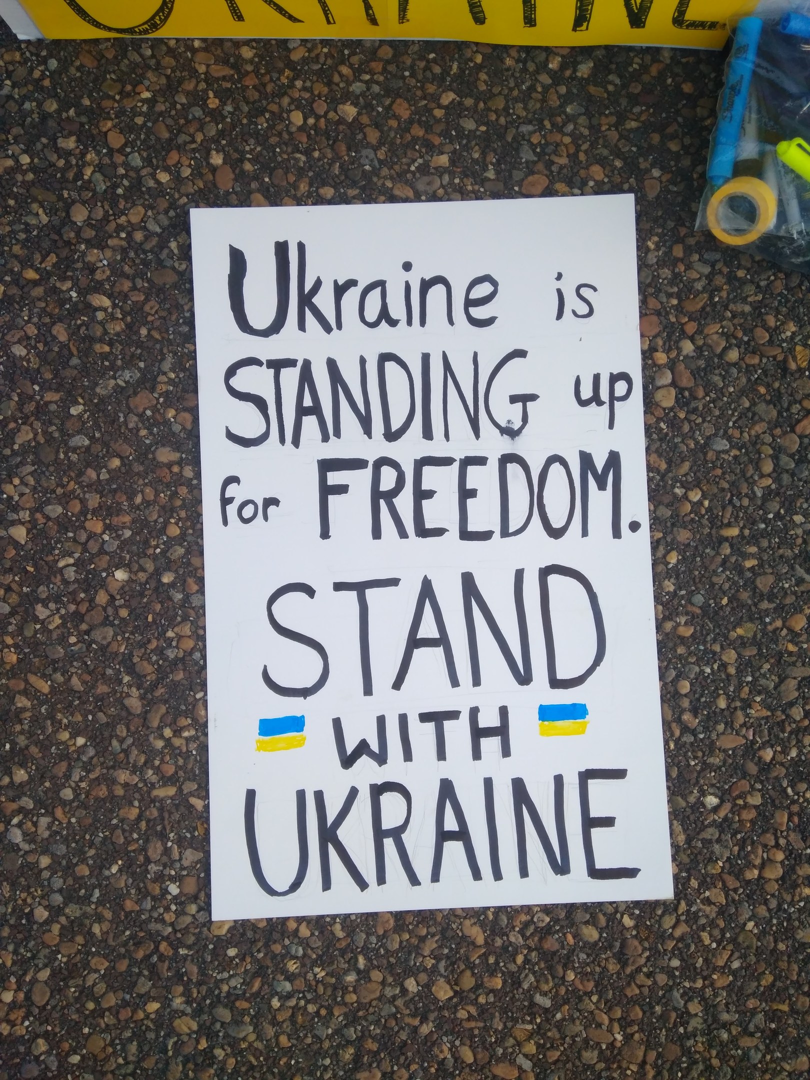 Ein weißer Zettel mit der Aufschrift "Ukraine steht für Freiheit, steht mit Ukraine" liegt vor einer gelben Fahne auf dem Boden. Rechts daneben befindet sich eine Plastikhülle mit verschiedenen Gegenständen.