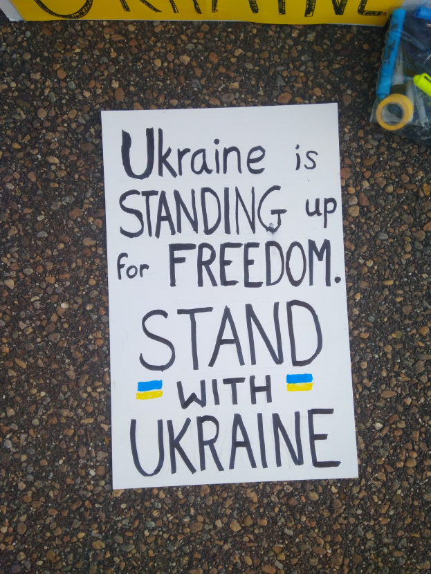 Ein weißer Zettel mit der Aufschrift "Ukraine steht für Freiheit, steht mit Ukraine" liegt vor einer gelben Fahne auf dem Boden. Rechts daneben befindet sich eine Plastikhülle mit verschiedenen Gegenständen.