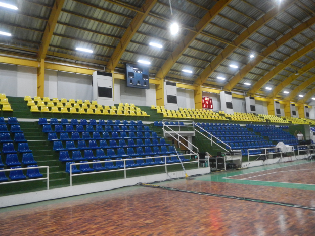 Eine geräumige Indoor-Sporthalle mit einem Holzboden, blauen und gelben Stühlen, Geländern, Treppen zum Sitzbereich, wandmontierten Bildschirmen, Deckenleuchten und einigen Anwesenden.
