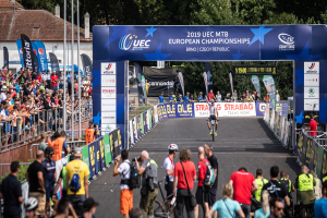 Eine Gruppe von Radfahrern fährt auf einer Straße entlang, die von Zuschauern gesäumt ist, mit einem Bogen darüber, auf dem "2019 UEC MTB Europameisterschaften" steht, und Gebäuden, Bäumen und einem klaren blauen Himmel im Hintergrund.