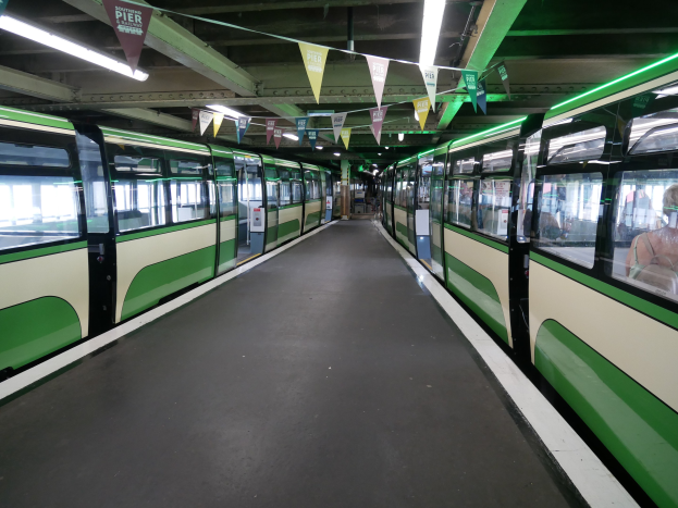 Ein grüner und weißer Zug mit Passagieren fährt an einem Bahnsteig vorbei unter einer Decke mit Lampen und dekorativen Fahnen an einem Münchner Bahnhof.