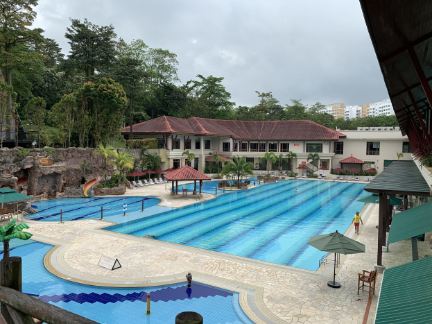Großes Schwimmbad im Ritz-Carlton, Kota Kinabalu, mit Menschen, Sonnenschirmen, Liegestühlen, Tischen, Pflanzen, Bäumen, Felsen, Gebäuden und einem Schuppen im Hintergrund.