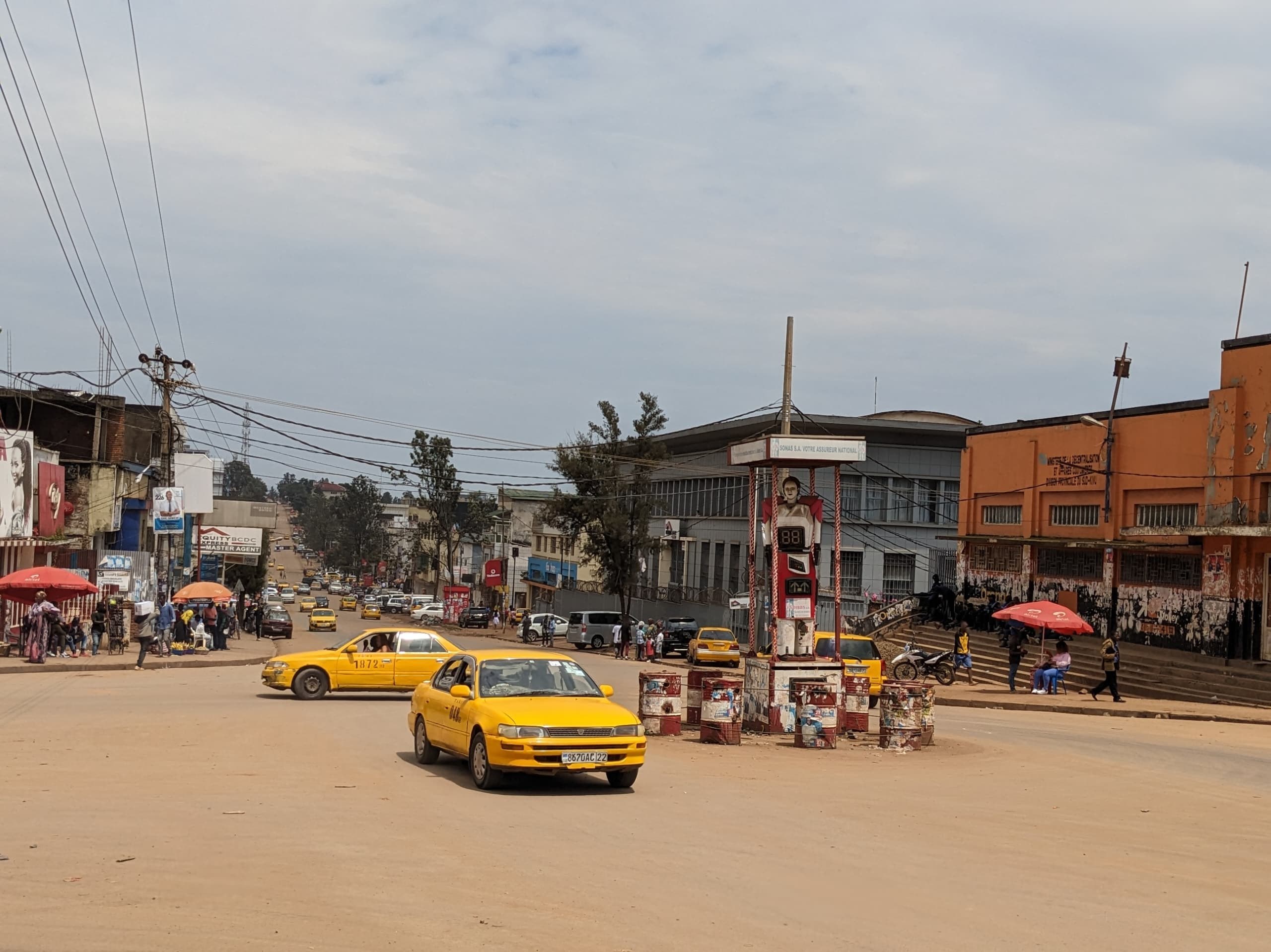 Ein gelbes Taxi fährt eine belebte Stadtstraße mit Gebäuden an den Seiten entlang, während Fußgänger mit Schirmen auf dem Gehweg darunter unter bewölktem Himmel gehen, mit Strommasten und Drähten im Hintergrund.