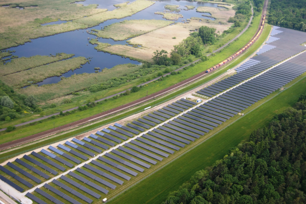 Luftaufnahme einer Solaränlage mit Panelen, einer Bahn auf einer Schiene, umgeben von Bäumen, Gras, Pflanzen und Wasser.