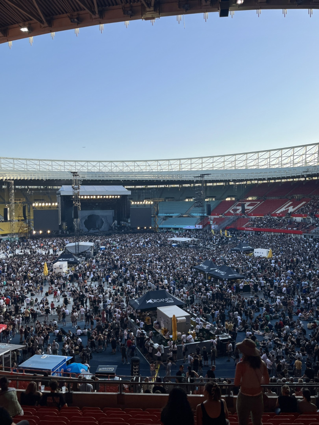 Eine große Menschenmenge sitzt und steht in einem stadionähnlichen Veranstaltungsort, mit Zelten, Geländern und Überdachung, unter einem überdachten Abschnitt, was auf ein Konzert in der O2 Arena in London hindeutet.