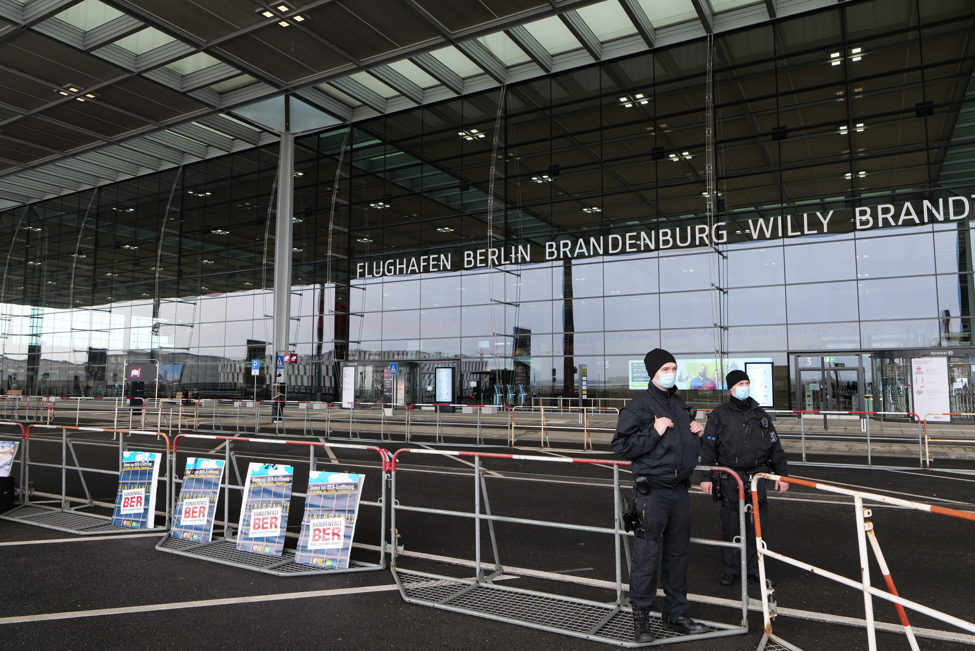 Zwei Polizisten in schwarzer Uniform stehen vor einem großen Glasgebäude mit Text, mit Absperrungen und Schildern im Vordergrund.