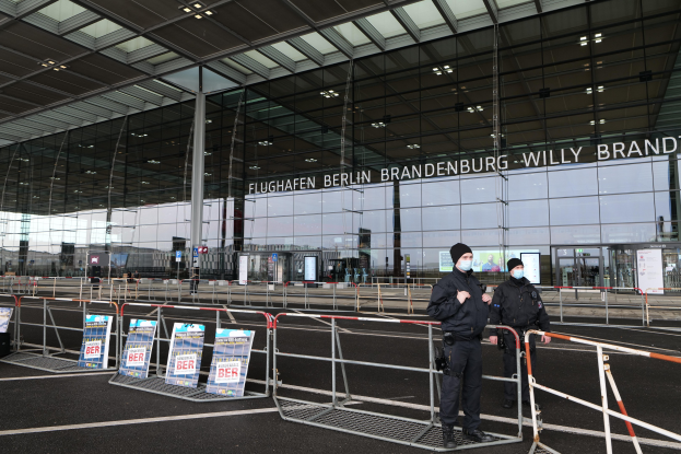 Zwei Polizisten in schwarzer Uniform stehen vor einem großen Glasgebäude mit Text, mit Absperrungen und Schildern im Vordergrund.