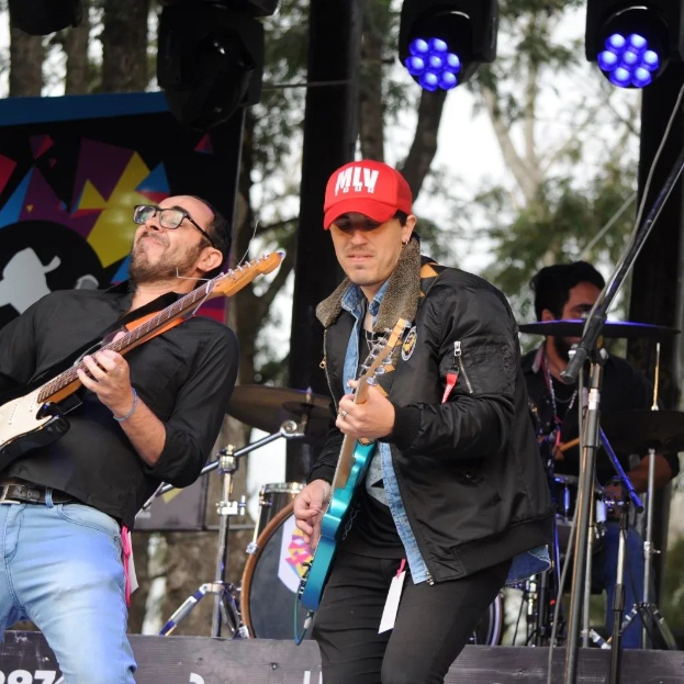 Zwei Männer auf der Bühne mit Gitarren, einer in schwarzer Jacke und blauen Hosen, der andere mit Brille und roter Mütze, während ein Schlagzeuger hinter ihnen sitzt und Mikrofone in der Nähe gegen einen Hintergrund von Bäumen unter einem klaren blauen Himmel stehen.