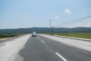 Ein Auto fahrt auf einer Hauptstra├če entlang, flankiert von Strommasten und Kabeln, mit saftig gr├╝nem Gras, B├Ąumen und H├Ąngen im Hintergrund unter einem klaren blauen Himmel.