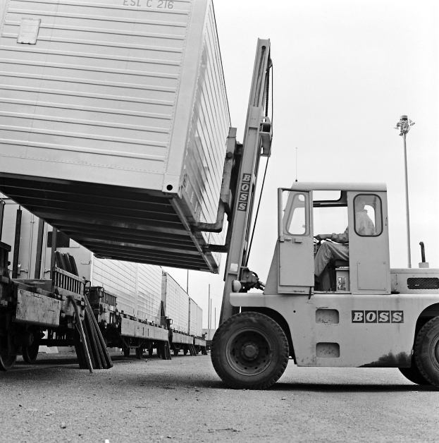 Ein Mann, der einen Gabelstapler mit einem Container auf dem Heck fährt, umgeben von anderen Fahrzeugen auf der Straße mit Pfählen und einem klaren Himmel im Hintergrund.