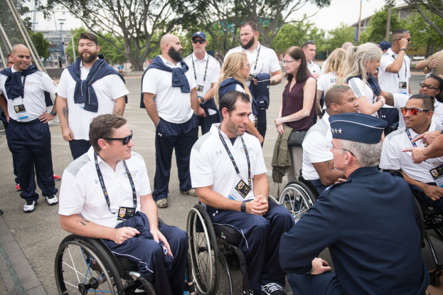 Eine Gruppe von Menschen im Rollstuhl unterhält sich im Freien, mit einer Kulisse aus Bäumen, Gebäuden und einem klaren blauen Himmel, einige tragen Mützen und Schutzbrillen, die auf eine Paralympische Teilnahme hinweisen.
