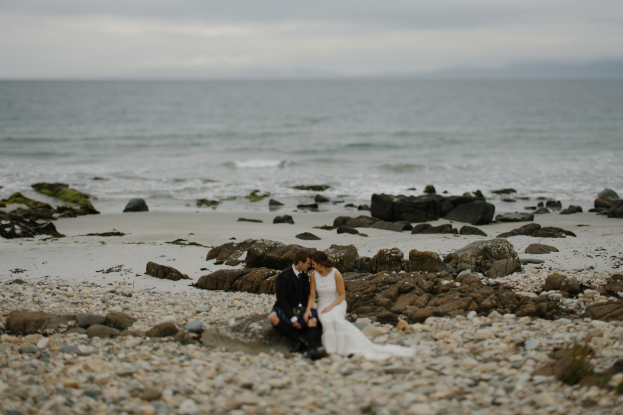 Braut und Bräutigam umarmen und küssen sich auf einem felsigen Strand mit dem Ozean und dem Himmel im Hintergrund.