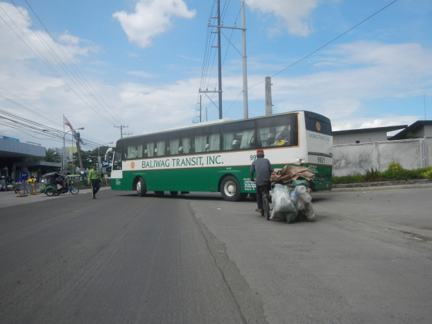 Grüner und weißer Bus fährt auf einer Straße mit Fußgängern, einem Radfahrer, Gebäuden, Bäumen und Strommasten im Hintergrund.