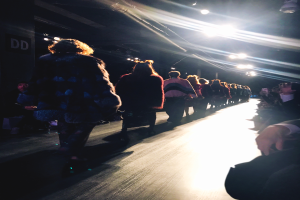 Eine Gruppe von Menschen läuft über einen hell beleuchteten Laufsteg bei einer Modenschau, mit Zuschauern auf beiden Seiten.